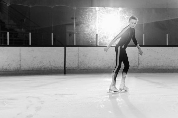 Black and white image of a girl figure skating on an ice rink indoors.